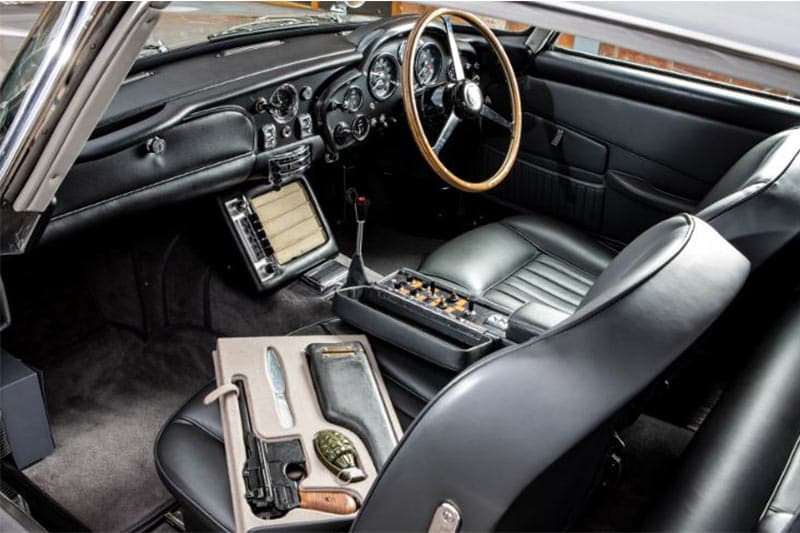The refurbished interiors of the Bond's Aston Martin DB5 look exquisite as if the car if fresh from the factory. Along with other gadgets, inside the cabin, this DB5 comes with a case that conceals some go-to weapons (fake). Notice the wooden steering wheel, all-black upholstery and analogue dials.