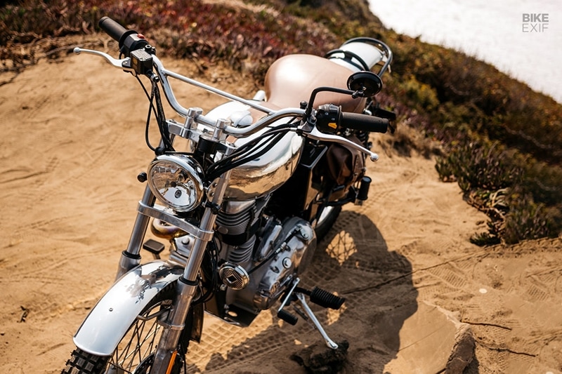 The crew also added a high-mounted single exhaust, and swapped the airbox for a pod filter. The Enfield’s original side ‘boxes’ (usually found under the seat) are gone—the right side now features an alloy side cover, and the left a leather bag that holds the electrics. Up in the cockpit is a new machined top yoke, with a wide set of scrambler bars. There’s a new headlight out front with a Motogadget speedo higher up, and both are mounted on custom-made brackets. (Source: Bikeexif)