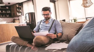 Modern bearded young businessman working on laptop computer while sitting on sofa at home, looking at some documents