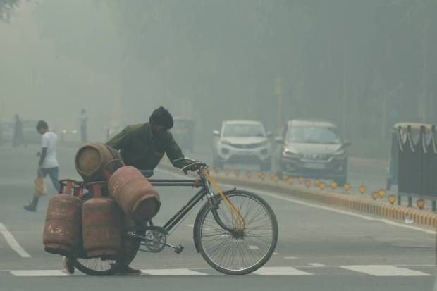 New Delhi: A worker carrying LPG cylinders on a bicycle crosses a road, shrouded in a thick layer of smog, in New Delhi. (PTI Photo)