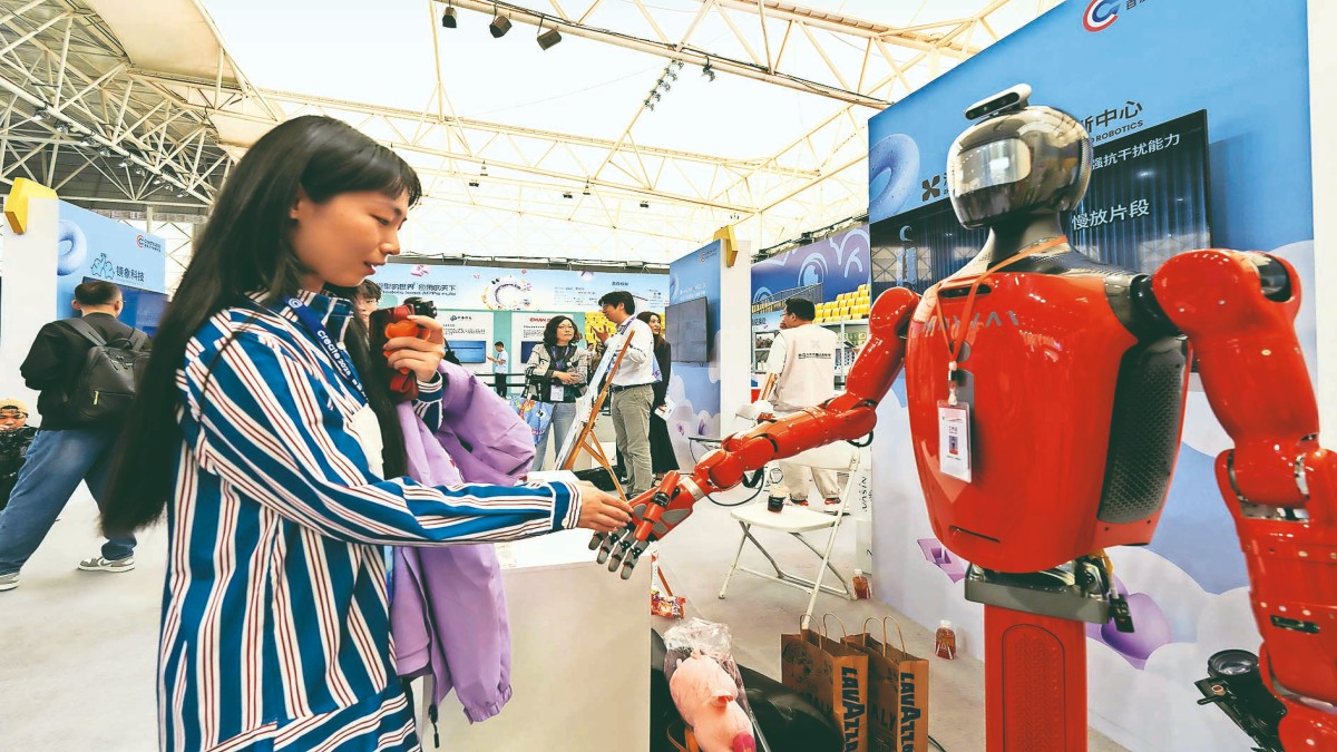 File image of a woman interacting with a humanoid robot during an AI developer conference in Wuhan, China. Even as we become more digitally interconnected, geopolitical rivalries remain fierce, often intensified by the very technologies that claim to transcend them. Photo credit: Bloomberg