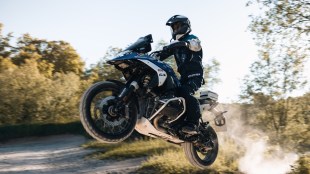 A motorcycle rider, fully equipped with a helmet, gloves, riding jacket, and knee guards, emphasizing the importance of essential safety gear for bikers and scooter riders