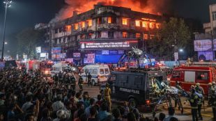 A high-stakes emergency scene in Nampally, Hyderabad, where a massive fire engulfs a multi-story building under a dark sky. Intense orange flames and thick black smoke pour from the windows. In the foreground, a large crowd of onlookers and emergency responders stand near Hyderabad Police vehicles. A specialized rescue robot is being deployed from a police van to assist in the operation. The atmosphere is chaotic and urgent, captured in a photorealistic news photography style.