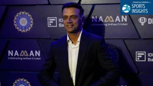 Rahul Dravid in a sharp navy blue suit standing in a modern Bangalore office complex, holding a tablet and looking towards a digital display, representing his role as a business mentor and investor.