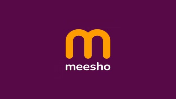 A delivery executive handling an e-commerce parcel with the distinct purple and orange color scheme of the Meesho brand outside an apartment complex.
